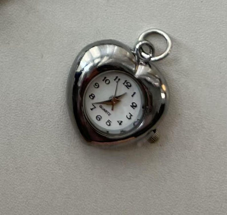 May include: A silver heart-shaped pendant watch with a white face and black numbers. The watch is quartz and has a small second hand.