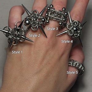 May include: A hand displays five silver rings. Styles 1, 2, and 4 feature spiked designs. Style 3 is a geometric structure of interconnected spheres and bars. Style 5 is a chainmail ring. The rings are labeled with the text "Style" and a number.