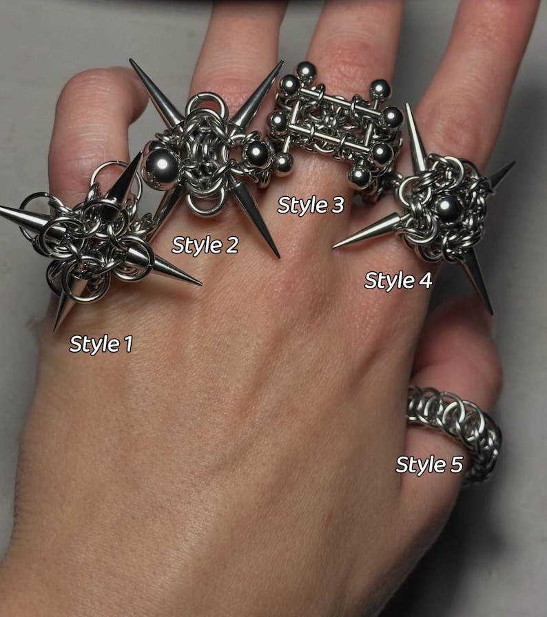 May include: A hand displays five silver rings. Styles 1, 2, and 4 feature spiked designs. Style 3 is a geometric structure of interconnected spheres and bars. Style 5 is a chainmail ring. The rings are labeled with the text "Style" and a number.