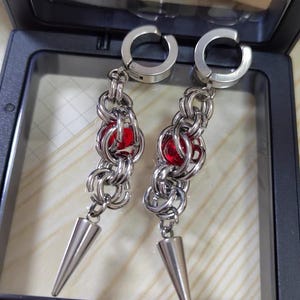 May include: A pair of silver earrings with red gem accents and a pointed tip. The earrings are made of metal chain links and have a hinged clasp.