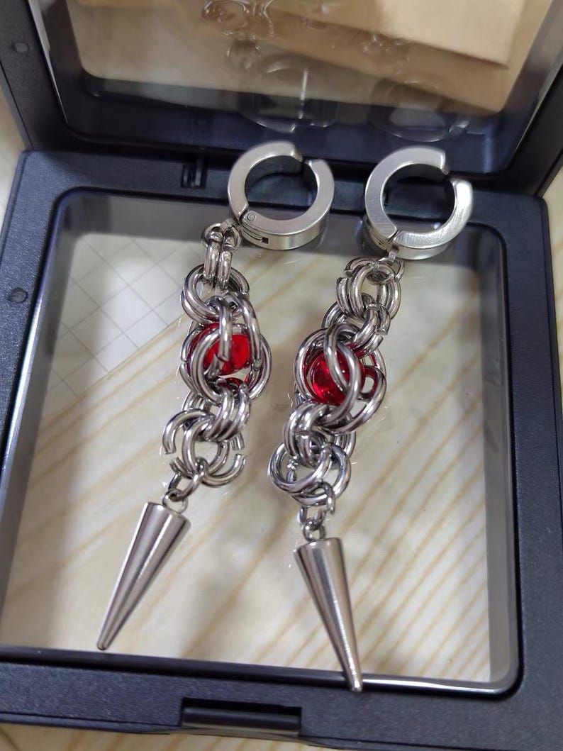 May include: A pair of silver earrings with red gem accents and a pointed tip. The earrings are made of metal chain links and have a hinged clasp.