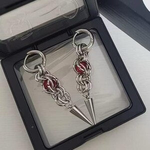 May include: A pair of silver earrings with red beads and pointed ends. The earrings are displayed in a black box.