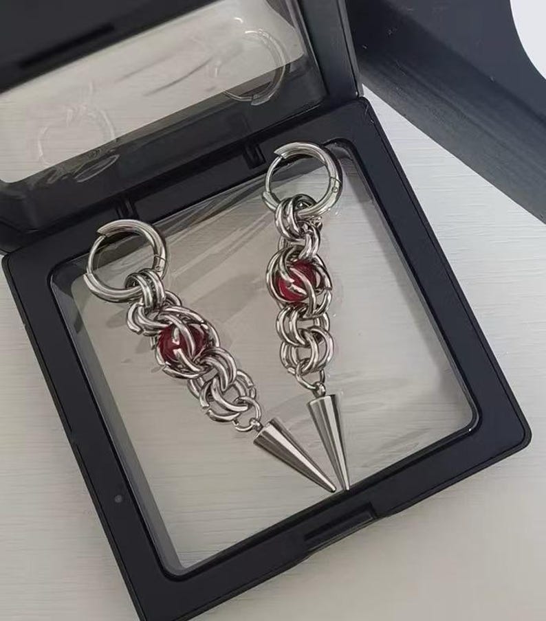 May include: A pair of silver earrings with red beads and pointed ends. The earrings are displayed in a black box.