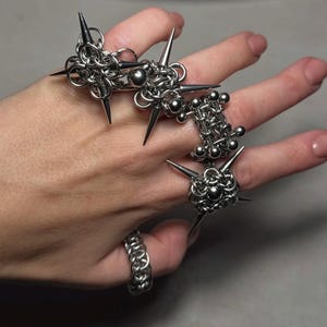 May include: A collection of silver-toned rings with a chainmail design. The rings feature sharp, conical spikes and small metal spheres. One ring is a simple chainmail band. The rings are displayed on a hand.