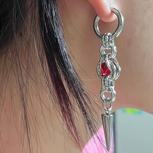 May include: Silver chain earring with a red gem and a pointed tip. The earring is hanging from a hoop.