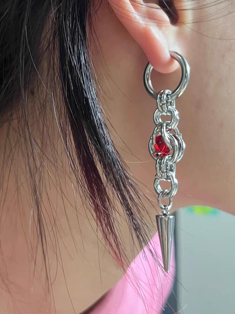 May include: Silver chain earring with a red gem and a pointed tip. The earring is hanging from a hoop.