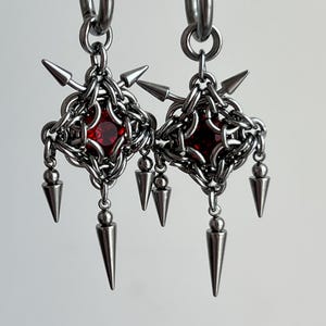 May include: A pair of silver earrings with a red gemstone in the center of each. The earrings are made of chainmail and have spikes hanging from the bottom.