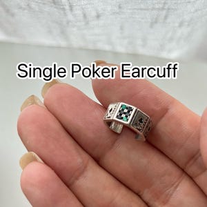 Puede incluir: Un earcuff de p&oacute;quer plateado &uacute;nico con un dise&ntilde;o rectangular. El earcuff presenta s&iacute;mbolos de naipes y piedras preciosas verdes y negras. El texto "Single Poker Earcuff" se muestra encima del earcuff.
