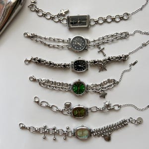 Puede incluir: Una colección de relojes de pulsera plateados con varias formas de esfera y estilos de cadena. Algunos relojes tienen esferas negras, mientras que otros tienen esferas verdes o claras. Cada reloj tiene dijes únicos, incluyendo cruces y estrellas.