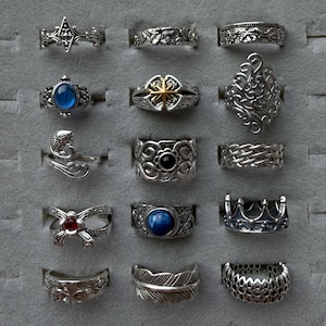 May include: A selection of silver rings with diverse designs, such as floral motifs, gemstones, and detailed filigree. Some rings feature a crown, a feather, and a fish. The rings are presented in a grey jewellery tray.