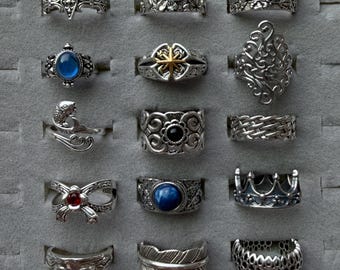 Peut inclure: Une collection de bagues en argent avec divers motifs, dont des motifs floraux, des pierres précieuses et des filigranes complexes. Certaines bagues présentent une couronne, une plume et un poisson. Les bagues sont exposées dans un présentoir à bijoux gris.