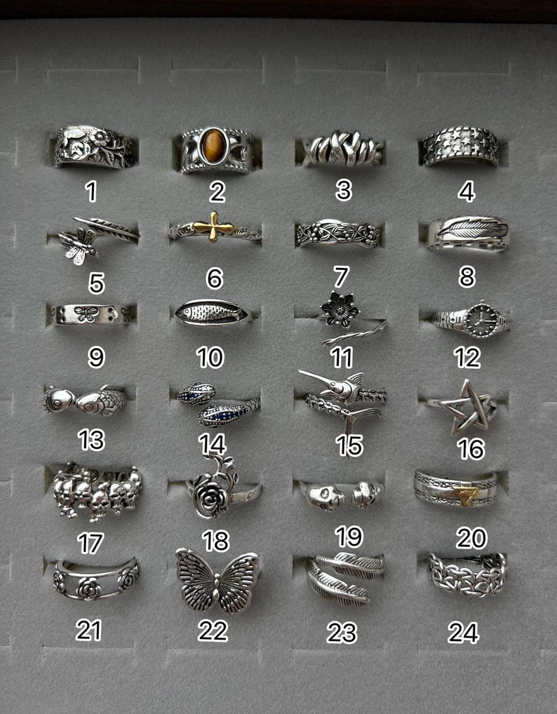 Puede incluir: Una colecci&oacute;n de veinticuatro anillos plateados exhibidos en un estuche gris. Los anillos presentan varios dise&ntilde;os, incluyendo patrones florales, animales y geom&eacute;tricos. Cada anillo est&aacute; numerado.