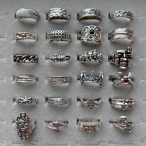 Op de afbeelding: Een verzameling zilveren ringen met diverse ontwerpen, waaronder getextureerde banden, bloempatronen en geometrische vormen. Sommige ringen hebben edelstenen of unieke motieven zoals schedels, kronen en veren. De ringen worden gepresenteerd op een grijze ondergrond.