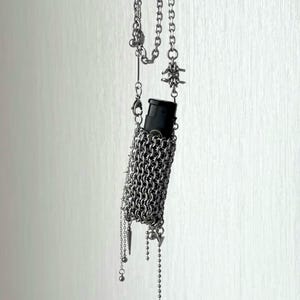 May include: A silver chain necklace with a black cylindrical object encased in a chainmail sleeve. The necklace features various chain styles, including barbed wire and decorative charms. The design has a gothic or punk aesthetic.
