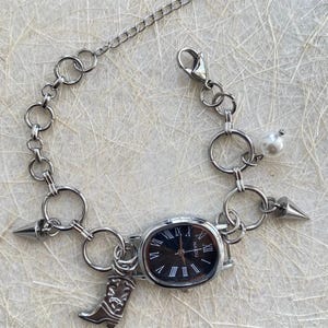 May include: A silver chain bracelet with a black watch face, a pearl charm, and a boot charm. The watch face has Roman numerals and the word "NADIU" printed on it.