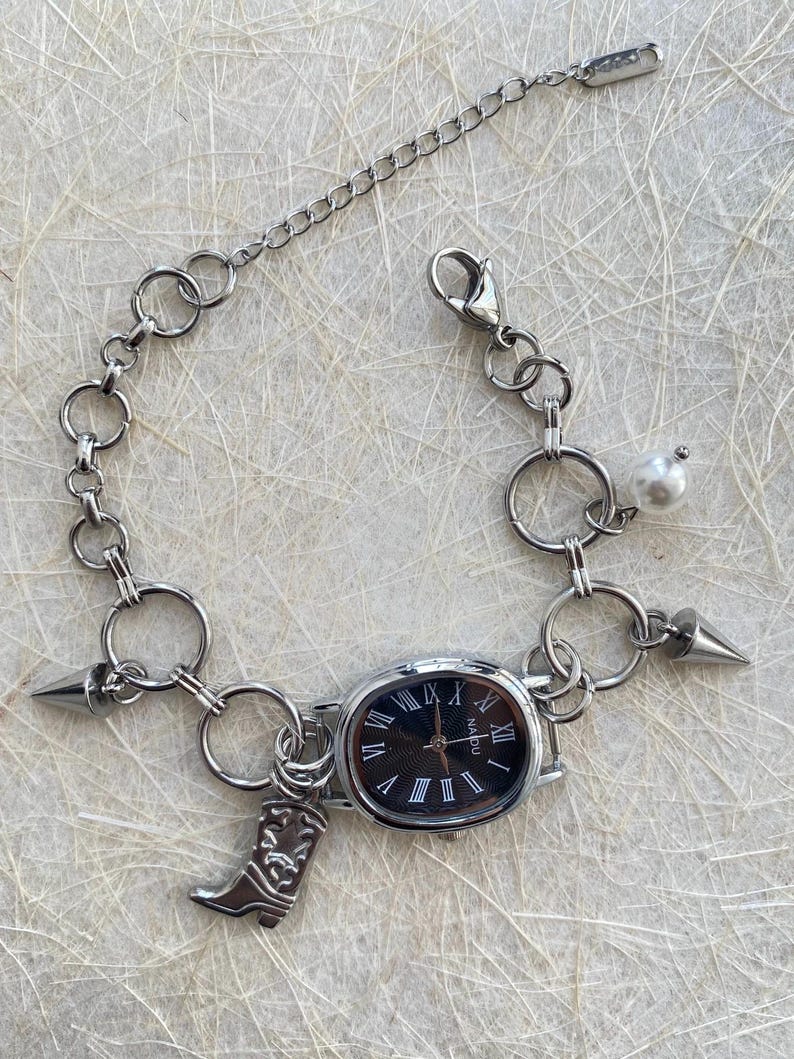 May include: A silver chain bracelet with a black watch face, a pearl charm, and a boot charm. The watch face has Roman numerals and the word "NADIU" printed on it.