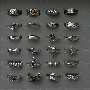 Puede incluir: Una colecci&oacute;n de anillos de plata con varios dise&ntilde;os, incluyendo una mariposa, una estrella, una pluma y una cruz. Los anillos se exhiben en una bandeja gris, mostrando los intrincados detalles y los diversos estilos de cada pieza.