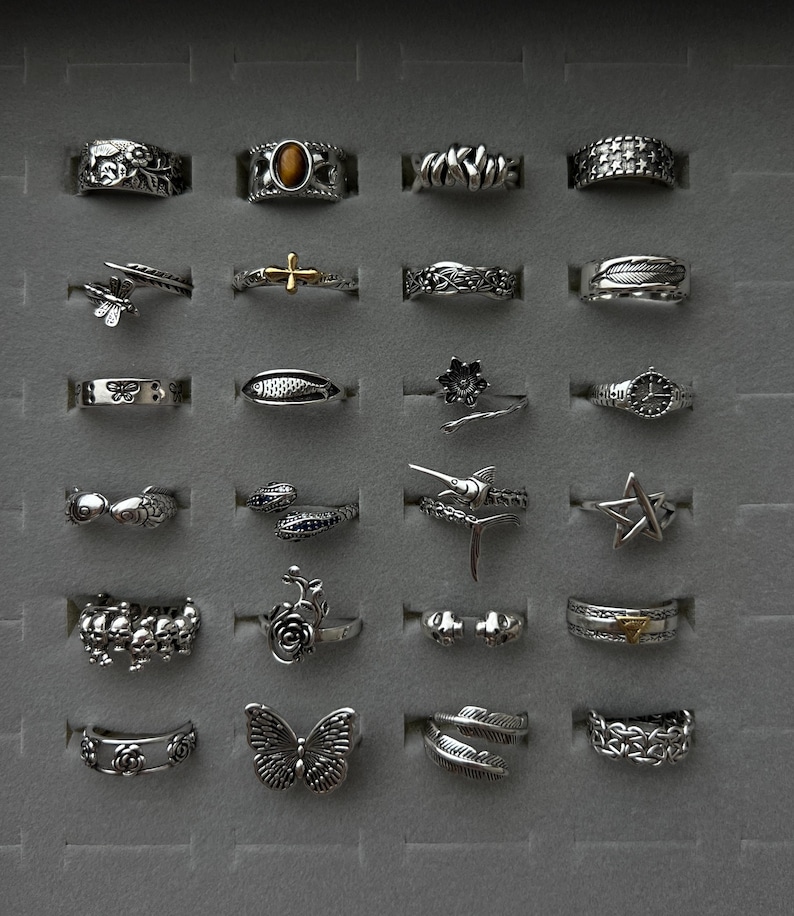 Puede incluir: Una colecci&oacute;n de anillos de plata con varios dise&ntilde;os, incluyendo una mariposa, una estrella, una pluma y una cruz. Los anillos se exhiben en una bandeja gris, mostrando los intrincados detalles y los diversos estilos de cada pieza.