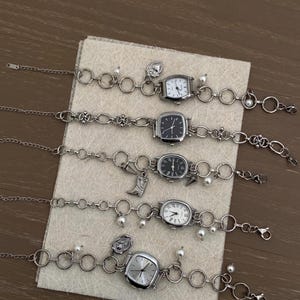 May include: Five silver charm bracelets with square watch faces. Each bracelet has a different charm, including a heart, a flower, a crescent moon, a cross, and a pearl.