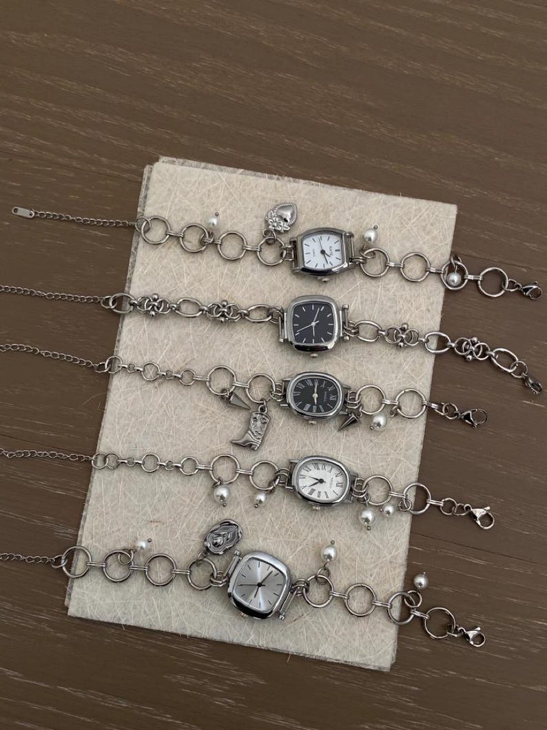 May include: Five silver charm bracelets with square watch faces. Each bracelet has a different charm, including a heart, a flower, a crescent moon, a cross, and a pearl.