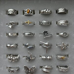 Puede incluir: Una colecci&oacute;n de anillos de plata exhibidos en una caja de joyer&iacute;a gris. Los anillos presentan varios dise&ntilde;os, incluyendo mariposas, flores, estrellas y patrones geom&eacute;tricos. Algunos anillos tienen detalles de piedras preciosas, y la colecci&oacute;n muestra una variedad de estilos y tama&ntilde;os.
