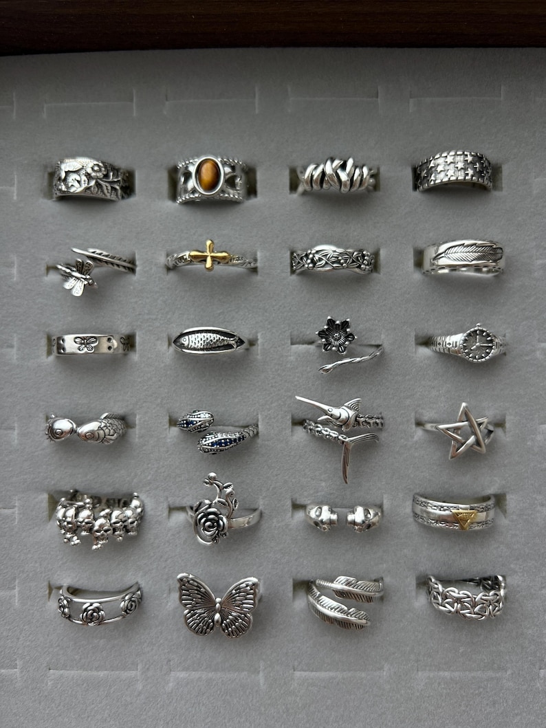 Puede incluir: Una colecci&oacute;n de anillos de plata exhibidos en una caja de joyer&iacute;a gris. Los anillos presentan varios dise&ntilde;os, incluyendo mariposas, flores, estrellas y patrones geom&eacute;tricos. Algunos anillos tienen detalles de piedras preciosas, y la colecci&oacute;n muestra una variedad de estilos y tama&ntilde;os.