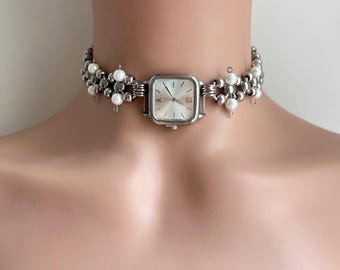 May include: A silver choker necklace with a square watch face and white pearl accents.