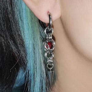 May include: Silver chain earrings with a red bead and a pointed tip. The earrings are hanging from a person's ear.
