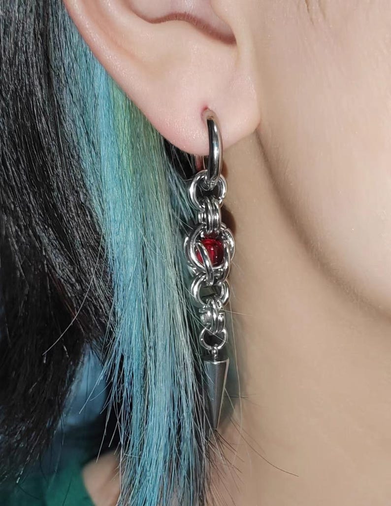 May include: Silver chain earrings with a red bead and a pointed tip. The earrings are hanging from a person's ear.