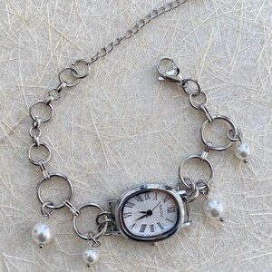 May include: A silver watch with a white face and Roman numeral markings. The watch is attached to a silver chain bracelet with multiple silver rings and white pearl charms.