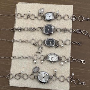 Puede incluir: Cinco pulseras de plata con diferentes esferas de reloj y dijes. Los dijes incluyen un corazón, una bota, una flor y una perla. Los relojes tienen correas plateadas y esferas blancas o negras.