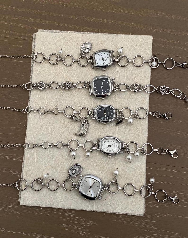 May include: Five silver charm bracelets with different watch faces and charms. The charms include a heart, a boot, a flower, and a pearl. The watches have silver bands and white or black faces.