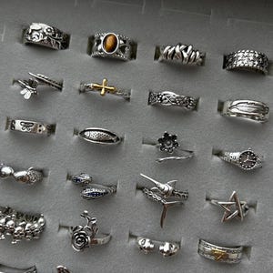 Puede incluir: Una colecci&oacute;n de anillos de plata con varios dise&ntilde;os, incluyendo patrones florales, animales y geom&eacute;tricos. Algunos anillos presentan piedras preciosas, mientras que otros tienen detalles intrincados como plumas, estrellas y cruces. Los anillos se exhiben en una bandeja de joyer&iacute;a gris.