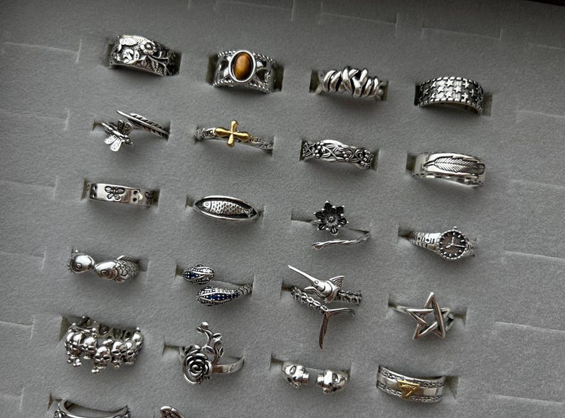 Puede incluir: Una colecci&oacute;n de anillos de plata con varios dise&ntilde;os, incluyendo patrones florales, animales y geom&eacute;tricos. Algunos anillos presentan piedras preciosas, mientras que otros tienen detalles intrincados como plumas, estrellas y cruces. Los anillos se exhiben en una bandeja de joyer&iacute;a gris.