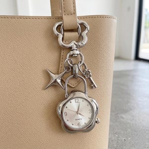 Könnte beinhalten: Ein silberfarbener Quarz-Uhrenanhänger mit blütenförmigem Zifferblatt, befestigt an einer beigefarbenen Handtasche. Der Anhänger enthält einen Stern, einen Schlüssel und einen blütenförmigen Verschluss. Das Zifferblatt zeigt die Zahlen 12, 3, 6 und 9.