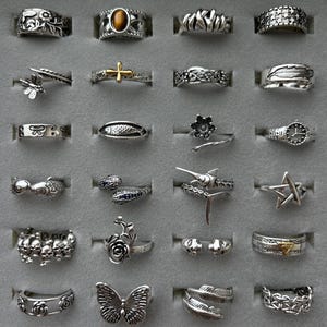 Op de afbeelding: Een verzameling zilveren ringen met diverse ontwerpen, waaronder een vlinder, een ster, een kruis en bloemmotieven. De ringen worden gepresenteerd in een grijze etui, waarbij de ingewikkelde details en gevarieerde stijlen worden benadrukt.