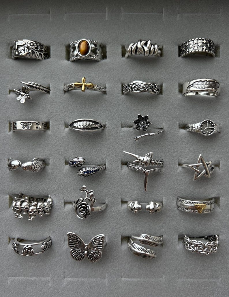 Puede incluir: Una colecci&oacute;n de anillos de plata con varios dise&ntilde;os, incluyendo una mariposa, una estrella, una cruz y motivos florales. Los anillos se exhiben en un estuche gris, mostrando los intrincados detalles y los diversos estilos.