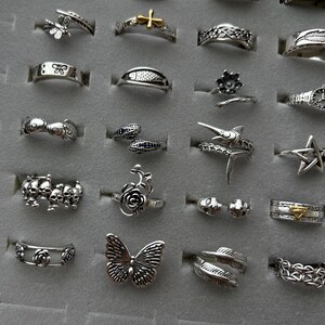 Puede incluir: Una colecci&oacute;n de anillos de plata con varios dise&ntilde;os, incluyendo mariposas, cruces, flores y calaveras. Los anillos se exhiben sobre una superficie gris, mostrando los detalles y estilos intrincados.
