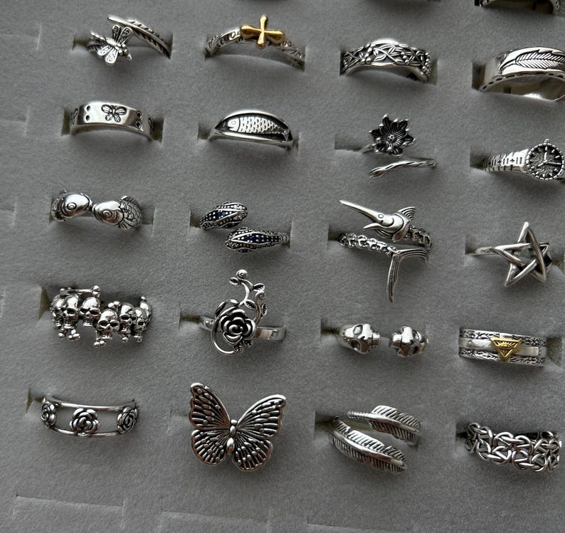 Puede incluir: Una colecci&oacute;n de anillos de plata con varios dise&ntilde;os, incluyendo mariposas, cruces, flores y calaveras. Los anillos se exhiben sobre una superficie gris, mostrando los detalles y estilos intrincados.