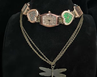 Könnte beinhalten: Eine roségoldene Halskette mit einem Libellenanhänger und einem Mehrfachuhren-Choker. Der Choker hat vier Uhren mit herzförmigen Zifferblättern, zwei mit schwarzen und zwei mit grünen Zifferblättern.