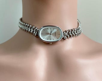 May include: A silver chain watch with an oval-shaped face. The watch face has a silver background with black markings and the word "NAIDU" printed on it.