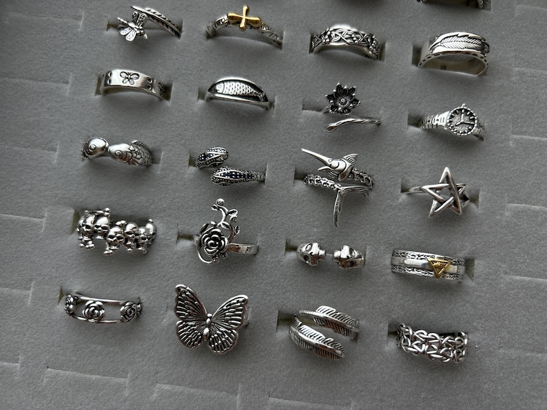 Puede incluir: Una colecci&oacute;n de anillos de plata con diversos dise&ntilde;os, incluyendo mariposas, flores y patrones geom&eacute;tricos. Los anillos se exhiben en una bandeja de joyer&iacute;a gris, mostrando sus intrincados detalles y estilos.