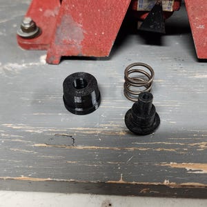Pode incluir: Close-up de três peças de plástico preto para uma prensa de recarga. As peças incluem uma mola, um botão pequeno e um botão maior com um furo rosqueado.