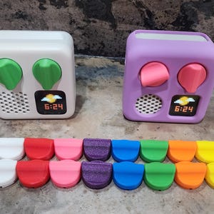 Könnte beinhalten: Zwei weiße und violette Wecker mit digitalen Displays, die 6:24 anzeigen. Die Wecker haben jeweils zwei Knöpfe, und es gibt 16 Ersatzknöpfe in verschiedenen Farben: Schwarz, Weiß, Rot, Rosa, Lila, Blau, Grün, Orange und Gelb.