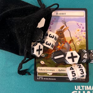 Könnte beinhalten: Eine schwarze Samt Tasche mit weißen Würfeln, die auf eine Magic: The Gathering Karte mit einer Kaninchenillustration fallen. Der Kartentext lautet "RABBIT" und "Token Creature - Rabbit".