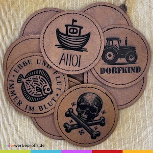 Könnte beinhalten: Sechs braune Lederpatches mit schwarzen Tintenmotiven. Die Motive beinhalten ein Boot mit dem Wort "AHOI", einen Traktor mit dem Wort "DORFKIND", einen Fisch mit den Worten "IMMER IM BLUT UND FLUT", einen Totenkopf und gekreuzte Knochen sowie zwei weitere Motive.