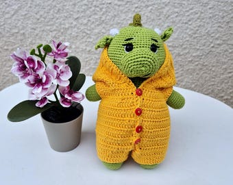 Peut inclure: Un dragon en crochet vert portant une combinaison en crochet jaune avec des boutons rouges. Le dragon est assis sur une surface blanche.