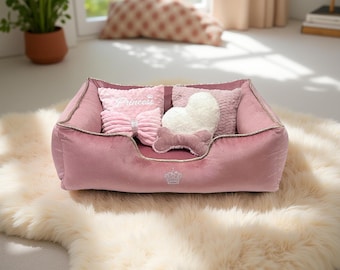 May include: A pink velvet pet bed with gold trim, filled with pillows. One pillow says "Princess" and has a bow. Another pillow is heart-shaped. The bed sits on a cream-colored, fluffy rug.