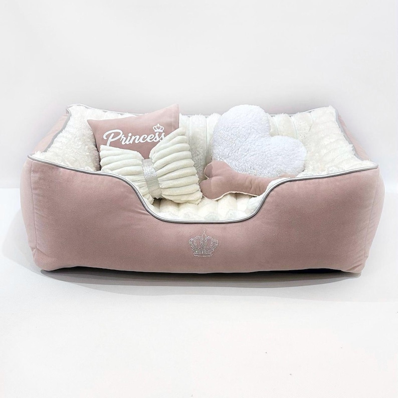 May include: A pink pet bed with a plush interior and a silver trim. The bed is decorated with pillows, including a heart-shaped pillow, a bow-shaped pillow, and a pillow with the word "Princess" on it. A crown is displayed on the front of the bed.