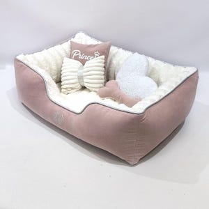 May include: A pink dog bed with a white interior and a gray trim. The bed is filled with pillows, including a bow-shaped pillow, a heart-shaped pillow, and a pillow with the word "Prince" embroidered on it. A pink bone-shaped toy is also included.